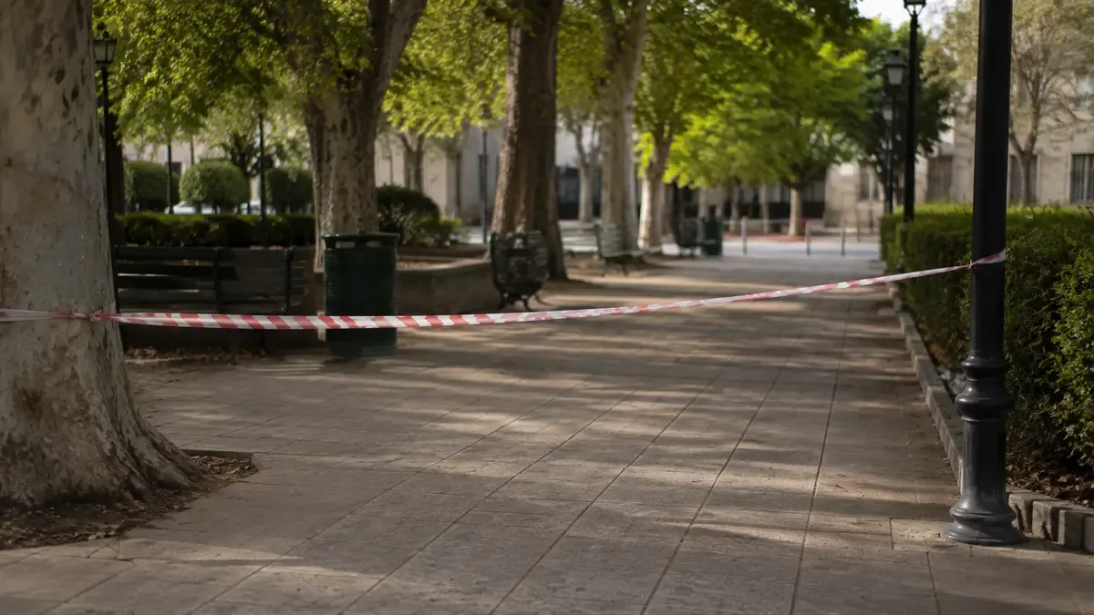 Cinta de seguridad lisa delimitando una vereda vacía en una plaza de barrio con mobiliario urbano, plano medio documental, perspectiva natural