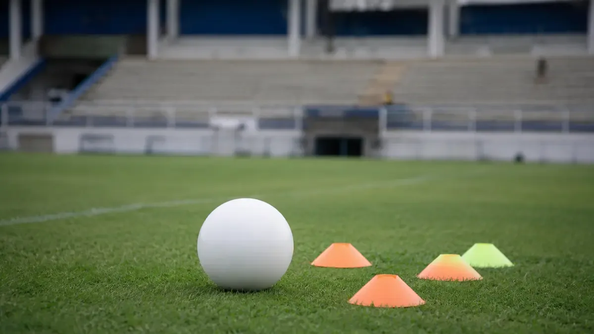 Una pelota lisa junto a conos de entrenamiento en un estadio local vacío, plano medio documental, perspectiva natural