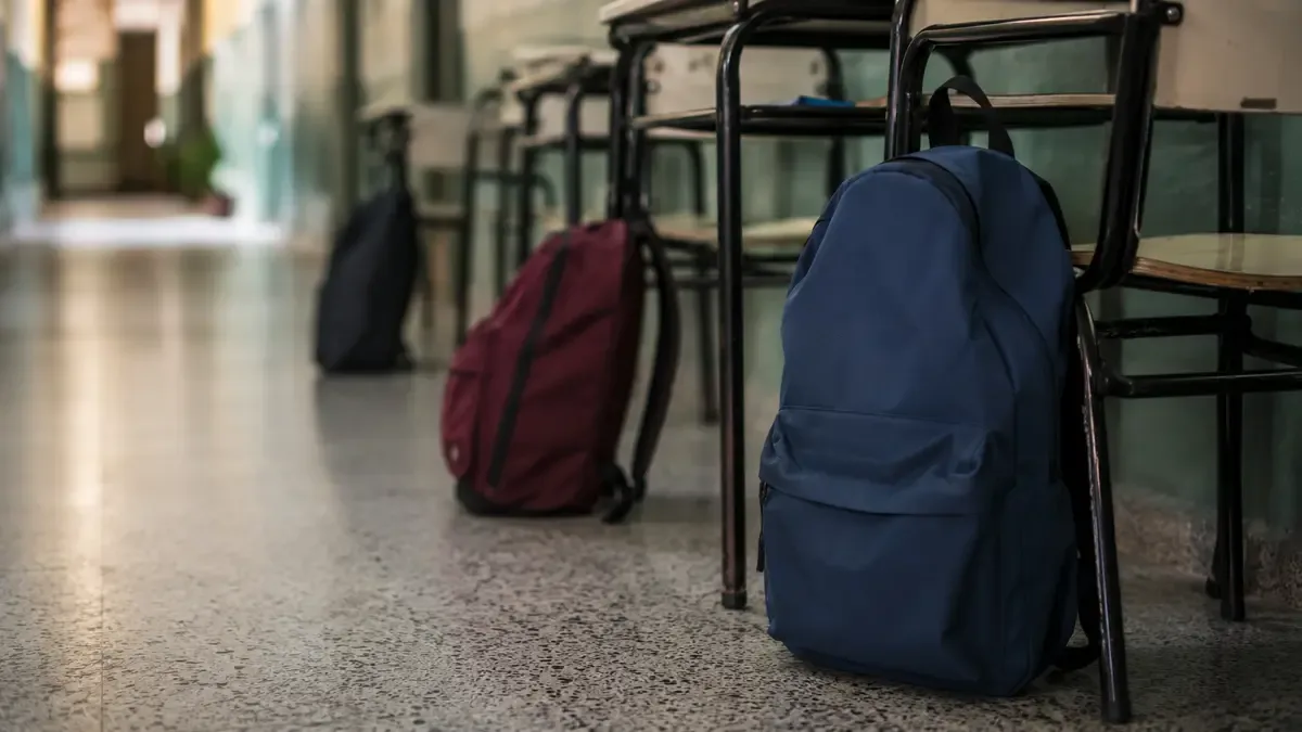 Mochilas lisas apoyadas junto a pupitres en un pasillo de escuela pública, vista en tres cuartos, profundidad de campo suave